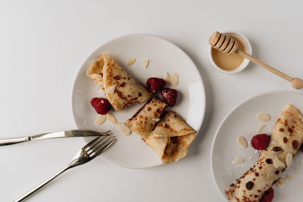 Draufsicht auf Crêpes mit Honig, Mandeln und Himbeeren auf einem weißen Teller.